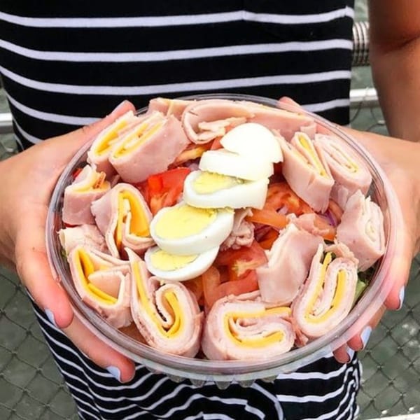 Hands holding a clear plastic bowl filled with ham and cheese roll-ups, tomatoes, and hard-boiled eggs.
