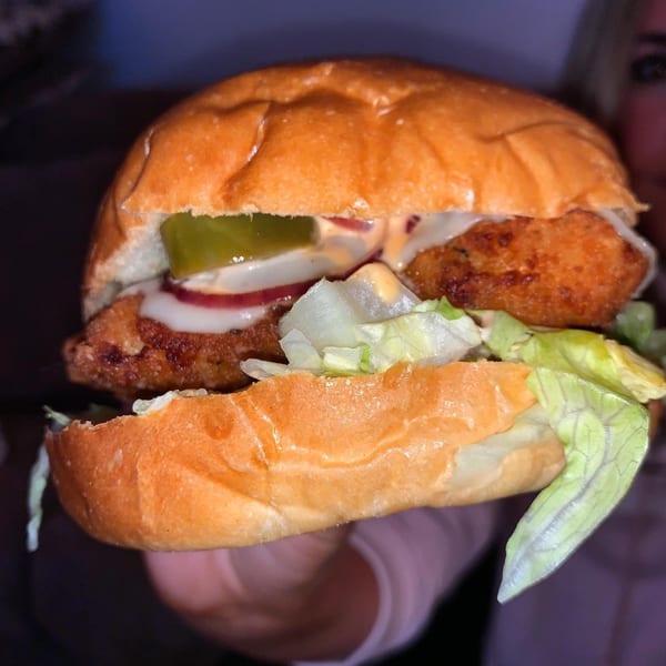 A hand holds a Grubbs Takeaway chicken burger with lettuce, pickles, onions, and sauce.