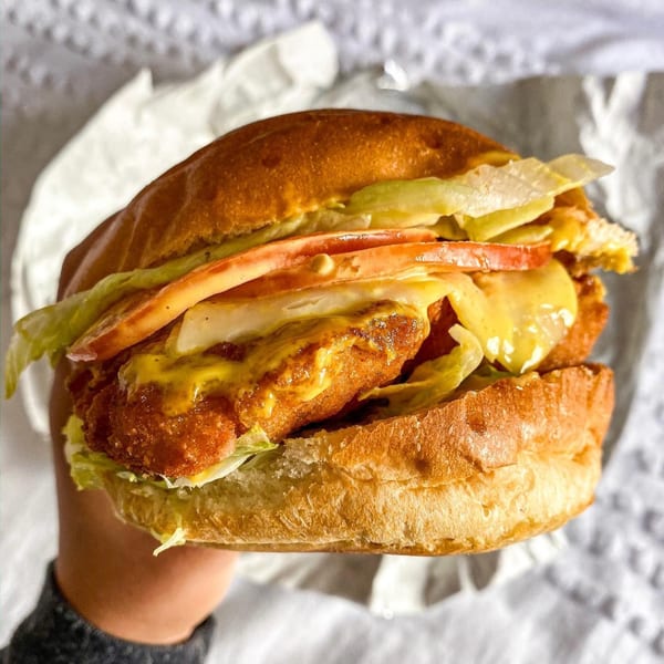 A hand holds a Grubbs Takeaway chicken burger with lettuce, tomato, cheese, and mustard.