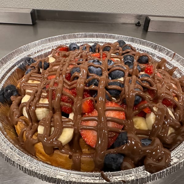 Waffle dessert in a foil pan, topped with strawberries, blueberries, bananas, and chocolate drizzle.