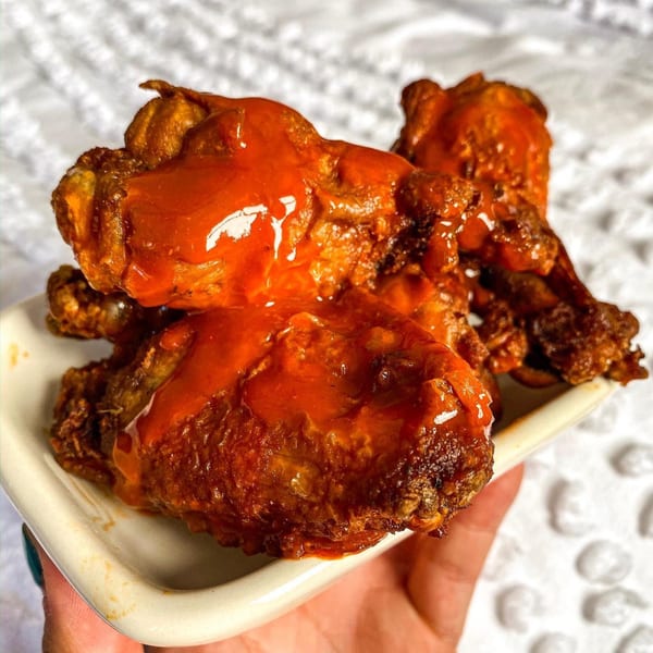 A hand holds a white dish filled with several crispy, saucy buffalo chicken wings from Grubbs Takeaway.
