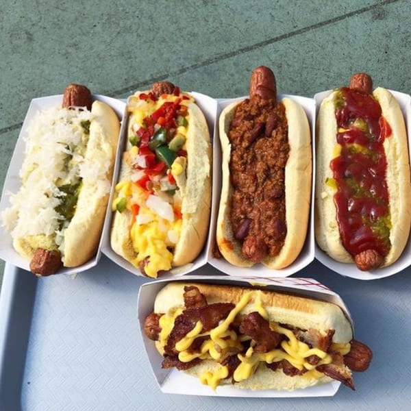 Five hot dogs with various toppings including chili, sauerkraut, relish, ketchup, mustard, and bacon.