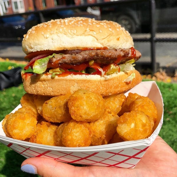 A Grubbs Takeaway burger with lettuce, tomato, and sauce, served with tater tots in a paper tray.